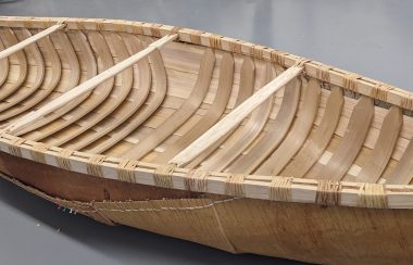 A plain wooden canoe sitting on a grey floor.
