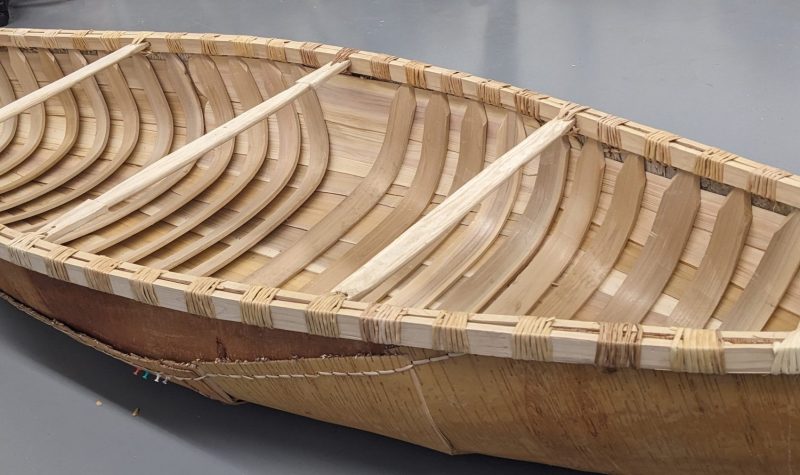 A plain wooden canoe sitting on a grey floor.
