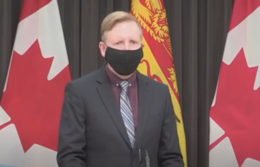 Dominic Cardy portant un masque noir devant un drapeau néo-brunswickois et canadien