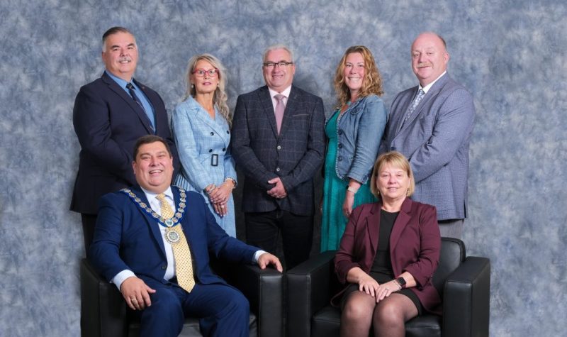 Corner Brook city council is in this group photo with a blue background. mayor Jim Parsons is sitting on the left and Councillor Charles Pender is standing directly behind him.