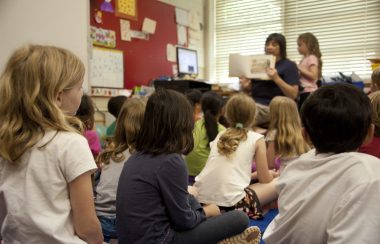 A teacher in the front showing a book to a room of children in a class room.