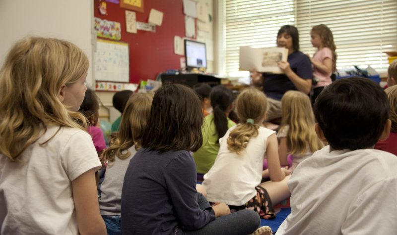 A teacher in the front showing a book to a room of children in a class room.