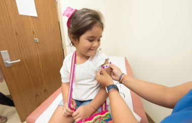 On aperçoit une petite fille souriante d'environ 5 ans qui vient de recevoir un vaccin et à qui on installe un pensement coloré