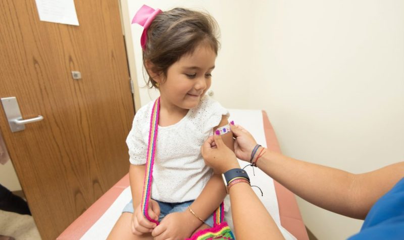 On aperçoit une petite fille souriante d'environ 5 ans qui vient de recevoir un vaccin et à qui on installe un pensement coloré