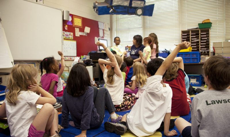 The photograph depicts a typical classroom scene, where an audience of school children were seated on the floor before a teacher at the front of the room, who was reading an illustrated storybook.