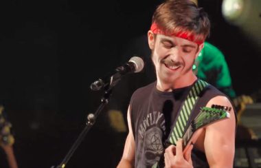 Un jeune guitariste et chanteur en action lors d'une spectacle. Il porte une camisole noire et un bandeau rouge sur la tête