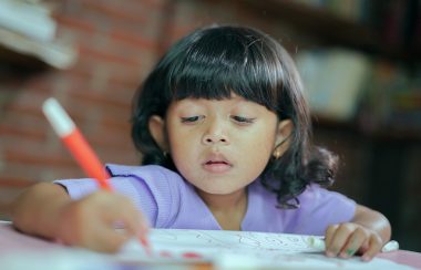 Une petite fille avec un chandail mauve qui colorie avec un stylo orange.