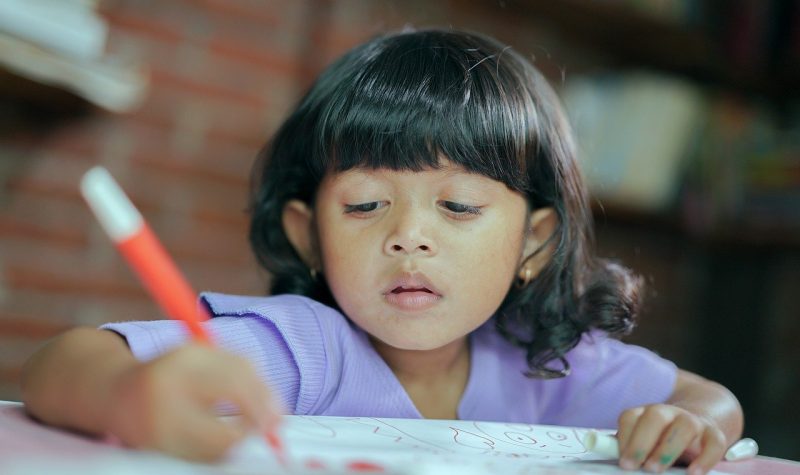 Une petite fille avec un chandail mauve qui colorie avec un stylo orange.