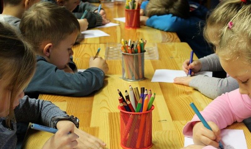 Des enfants sont assis à une table dans une garderie pour dessiner. On peut observer un pot avec des crayons de couleurs.