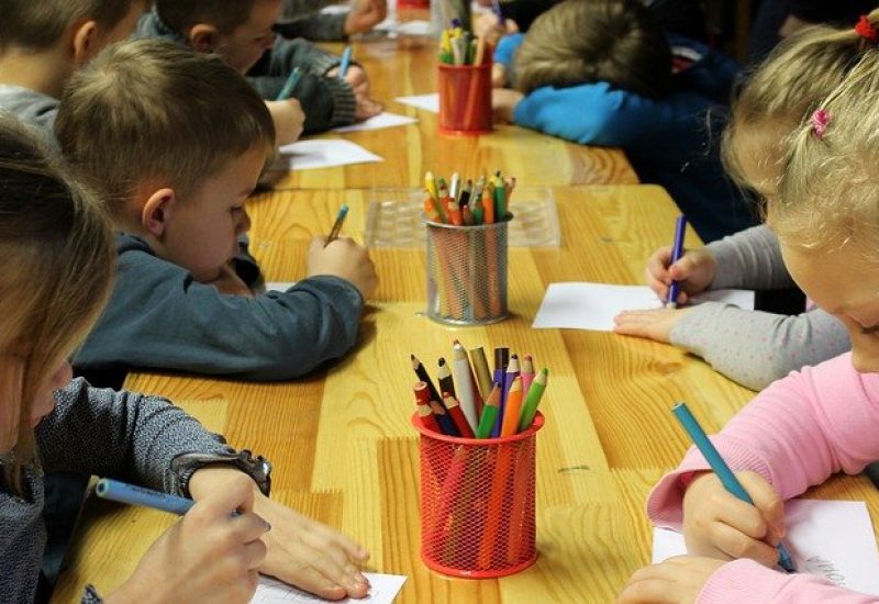 Des enfants sont assis à une table dans une garderie pour dessiner. On peut observer un pot avec des crayons de couleurs.