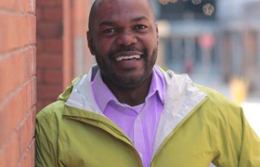 A person stands on a brick wall to their right as they wear a yellow jacket and purple collared shirt.