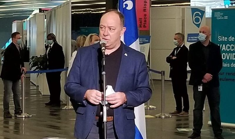 Le ministre christian dubé parle dans un micro au Palais des congrès de Montréal. Il est posé devant un drapeau du Québec. Il porte un veston bleu et un jean.