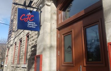 En haut à gauche, le logo rouge du centre communautaire Jacques-Cartier est inscris sur un panneau bleu. À la droite de ce panneau, la porte d'entrée du centre communautaire.