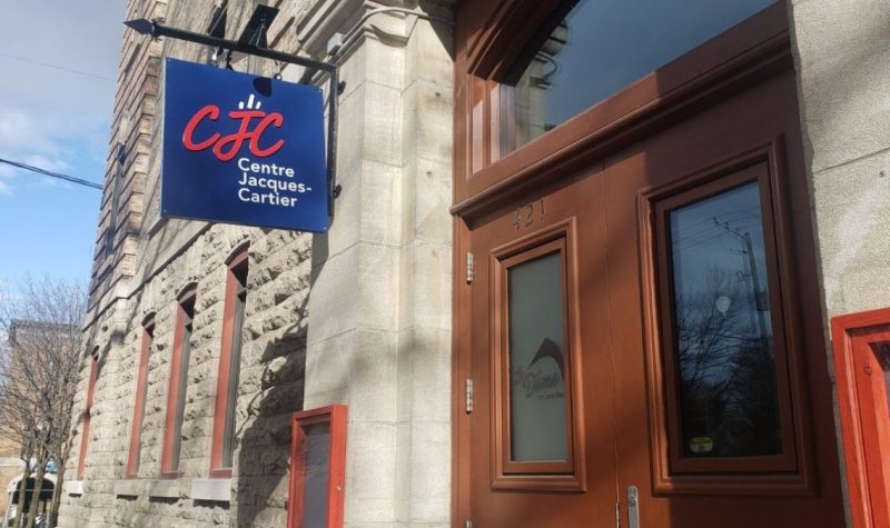 En haut à gauche, le logo rouge du centre communautaire Jacques-Cartier est inscris sur un panneau bleu. À la droite de ce panneau, la porte d'entrée du centre communautaire.