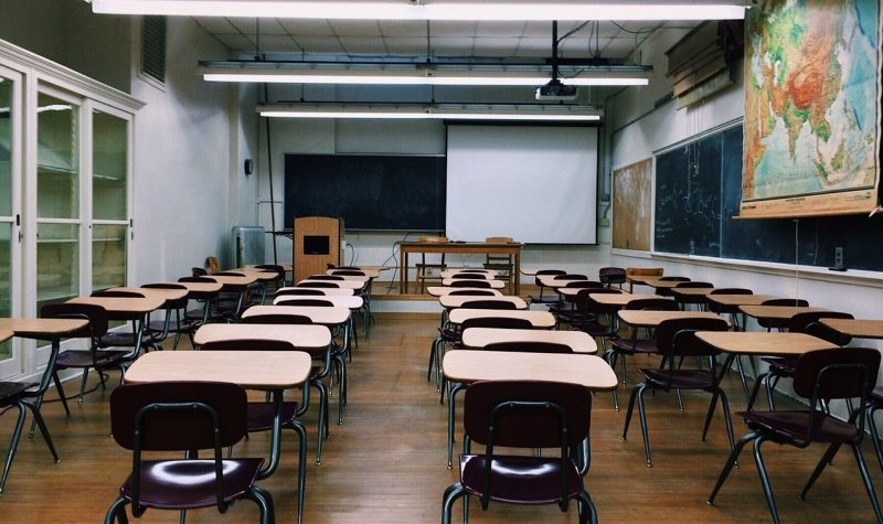 A classroom filled with a number of individual sized desks and chairs lined in rows. Black chalkboards can be seen on the side and front walls, and a world map can also be seen on the side wall.