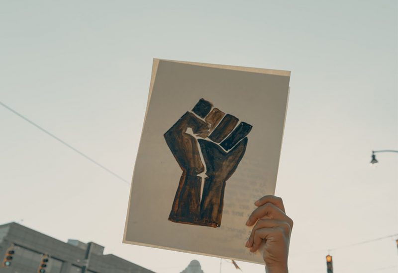 An image of a painting of a black fist representing Black Lives Matter. It is held up against a blue sky.