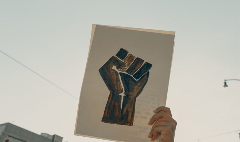 An image of a painting of a black fist representing Black Lives Matter. It is held up against a blue sky.