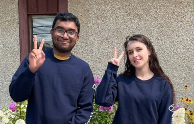 A man and woman wearing matching blue sweaters give a peace sign to the camera.