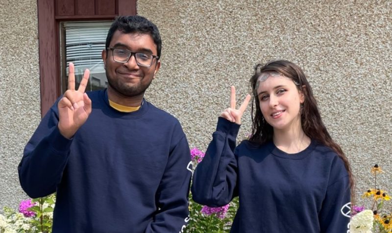 A man and woman wearing matching blue sweaters give a peace sign to the camera.
