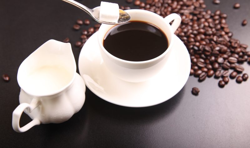 Une tasse de café blanche, a gauche un petit pot de lait, des grains de café sont disposés sur la table, une cuillère est prête à déposer du sucre dans la tasse.