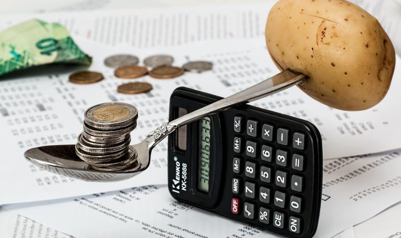 Sur une table jonchée de feuilles et de monnaie, une patate et des pièces de monnaie se tiennent en équilibre sur une calculatrice placée à la verticale.