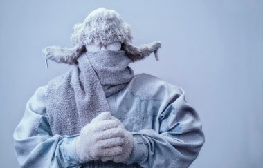 A frozen man with his hands clasped all bundled up in a fur trappers hat, scarf, parka and knit gloves, covered in snow and frost trying to stay warm on a very cold gray Winter's day.