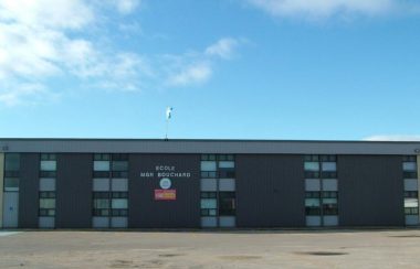 École primaire Mgr Bouchard, bâtisse grise foncée dans un stationnement vide avec un ciel bleu dégagé en arrière plan.