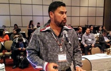 Six Nations Councilor Nathan Wright stands at a podium in front of a microphone. Audience Members look on in the background.