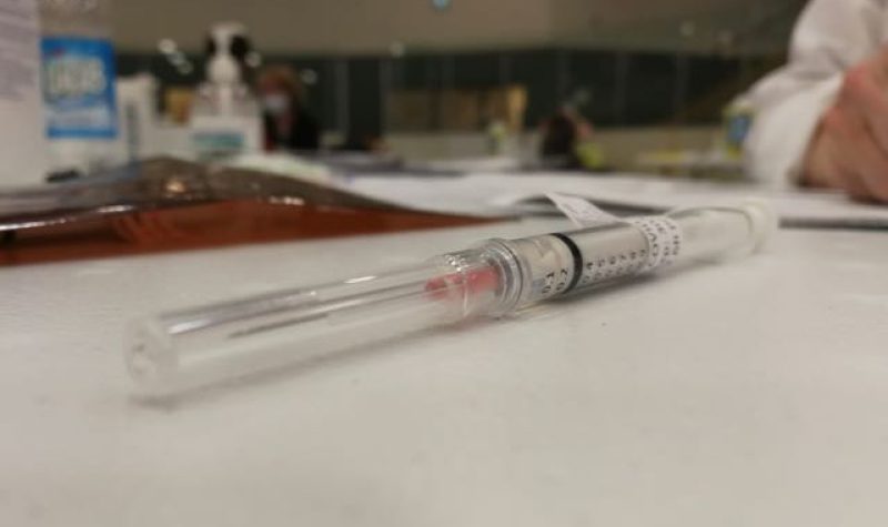 A capped syringe sits on a table with hand sanitizer and a person's elbow resting on the table in the background.