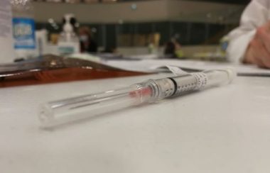 A needle containing a shot of Pfizer vaccine sits on a table, with a hand and paperwork in background.