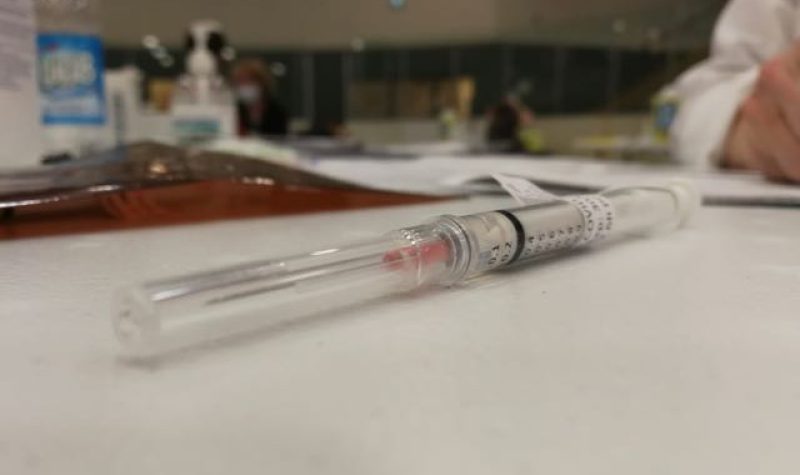 A needle containing a shot of Pfizer vaccine sits on a table, with a hand and paperwork in background.