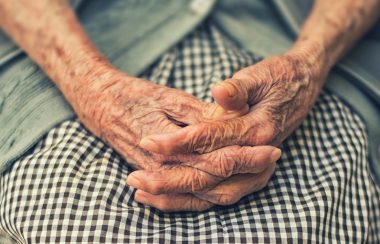 An elderly person folds their hands in their lap.