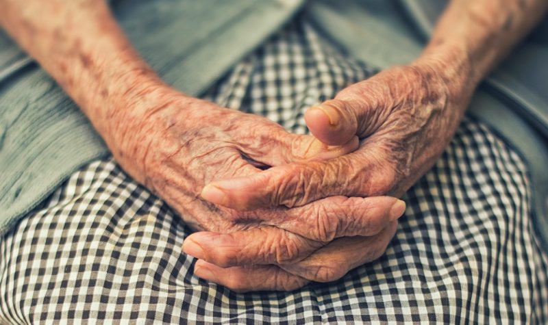 An elderly person folds their hands in their lap.