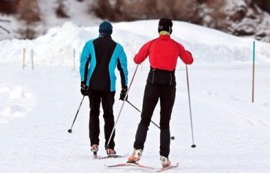 Deux skieurs de fond sur une piste. Le skieur à gauche porte un manteau noir et bleu. La skieuse de droite porte un manteau rouge. La piste est recouverte de neige.