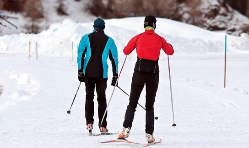Deux skieurs de fond sur une piste. Le skieur à gauche porte un manteau noir et bleu. La skieuse de droite porte un manteau rouge. La piste est recouverte de neige.