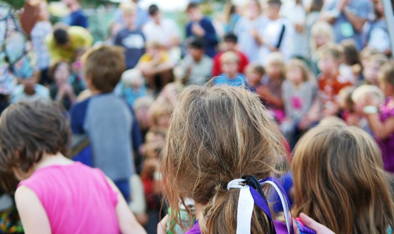 Crowd of children gathering in a spot. The back of a girls head is shown. Se is wearing a purple shirt and has a blue and white ribbon in her ponytail. Other children can be seen in the background.