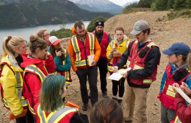 a group of people are standing in a circle around one man holding a clipboard. They are listening to him explain something and there is a river and mountains behind them.