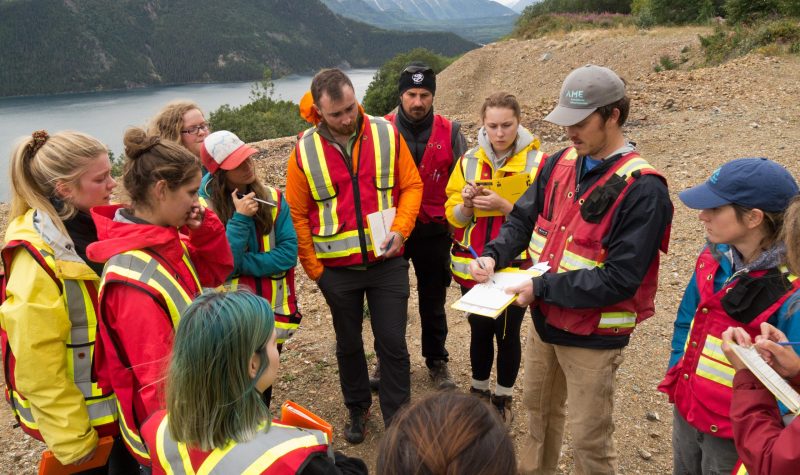 a group of people are standing in a circle around one man holding a clipboard. They are listening to him explain something and there is a river and mountains behind them.