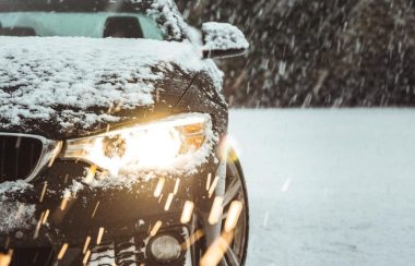 Une voiture sur une terrain enneigé. Les phares sont allumés et on peut voir que la photo à été prise alors que le ciel est sombre. Il tombe de la neige sur la voiture.