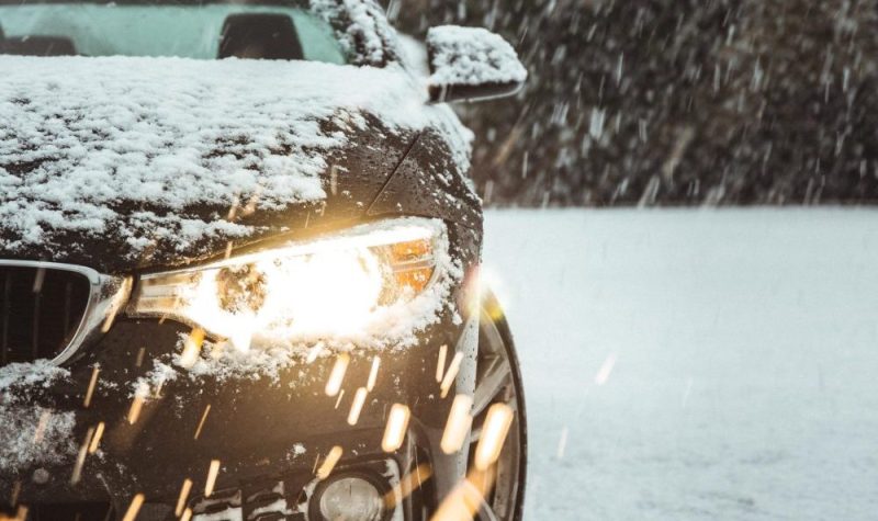 Une voiture sur une terrain enneigé. Les phares sont allumés et on peut voir que la photo à été prise alors que le ciel est sombre. Il tombe de la neige sur la voiture.