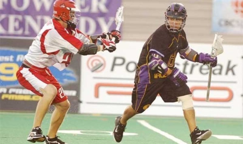 Two men playing field lacrosse. A man in the black uniform carries the ball against a man in a red and white uniform.