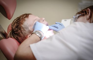 Une hygiéniste dentaire travaille, ses mains protégées par des gants bleus, dans la bouche d'un jeune patient allongé.
