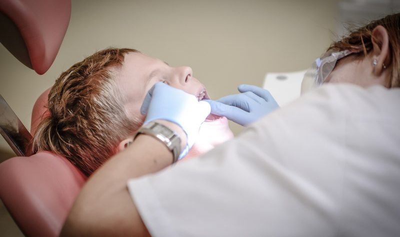 Une hygiéniste dentaire travaille, ses mains protégées par des gants bleus, dans la bouche d'un jeune patient allongé.