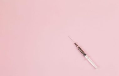 A single syringe sits on a pink background.