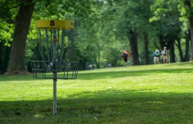 A disc golf basket sitting in a field with trees on both it's sides and a group of disc golfers throwing frisbees at the basket.