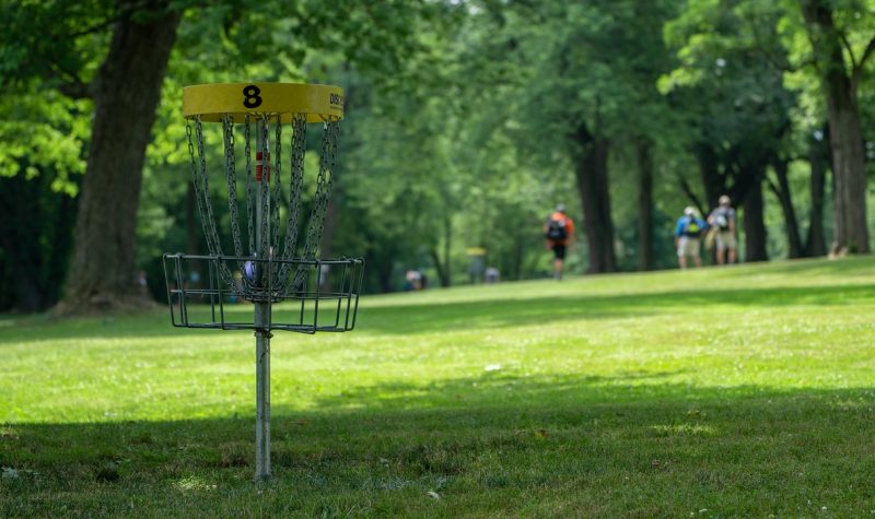 A disc golf basket sitting in a field with trees on both it's sides and a group of disc golfers throwing frisbees at the basket.
