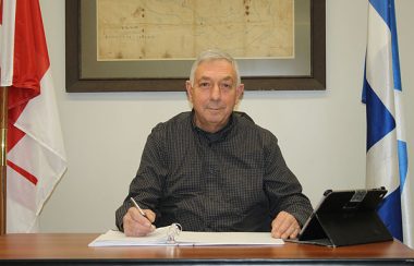 Chichester Mayor Donald Gagnon sits at his desk flanked by a Canadian and Quebec flag and wearing a dark shirt.