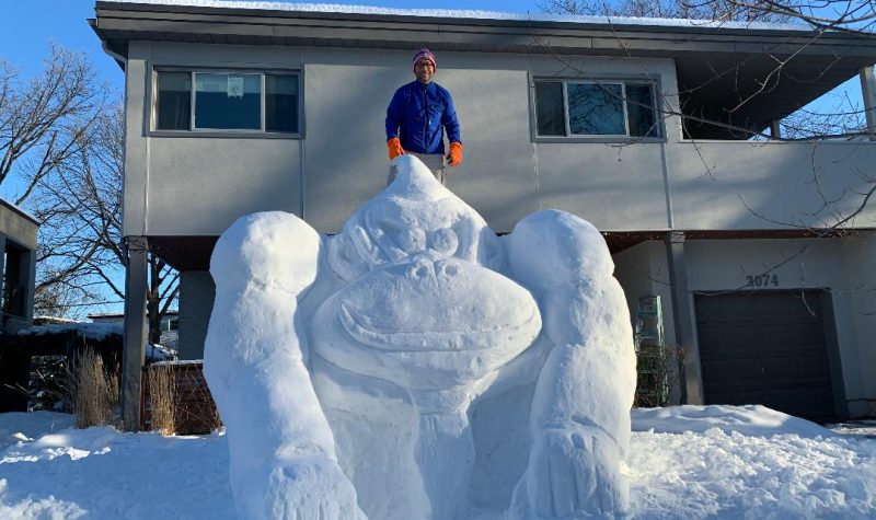 A 12-foot sculpture of Donkey Kong made out of snow is seen on someone's snowy front lawn, with a man standing at the top, wearing a blue winter coat and orange gloves.
