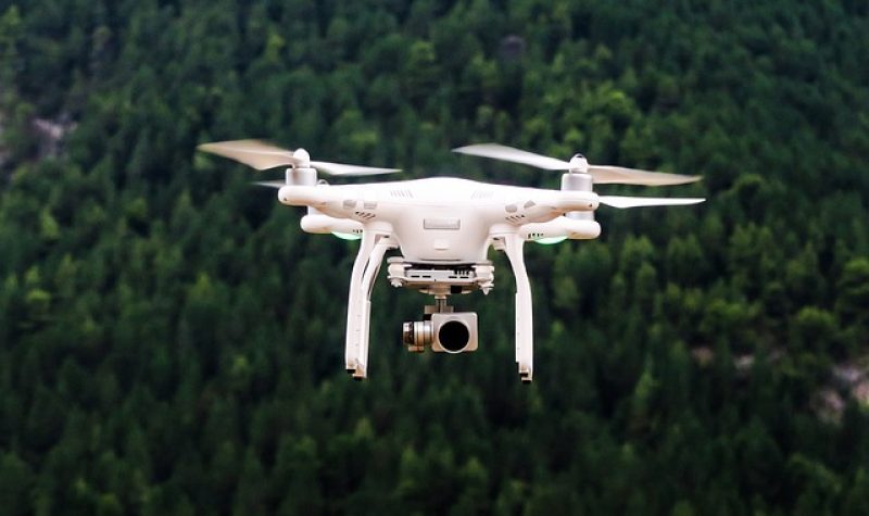 Un appareil volant blanc est au dessus d'une grande forêt. Il porte une caméra.
