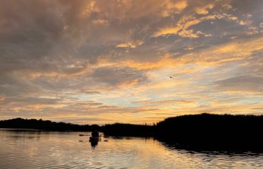 A kayaker can be seen paddling on water as a marsh sits in the background. The sky is filled with clouds and blue skies.